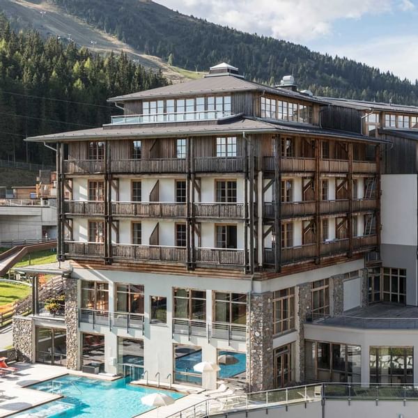 Modernes Berghotel mit Pool und vielen Fenstern, umgeben von einer bewaldeten Berglandschaft.