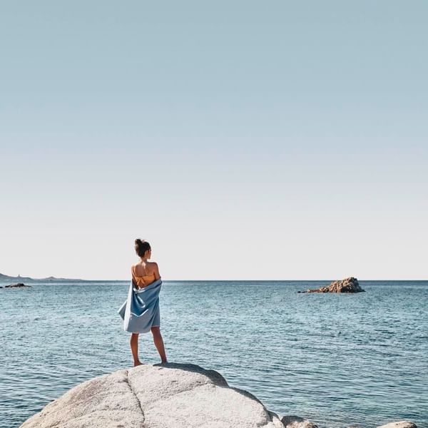 Frau steht auf einem Felsen am Meer und schaut hinaus, in ein Handtuch gewickelt.