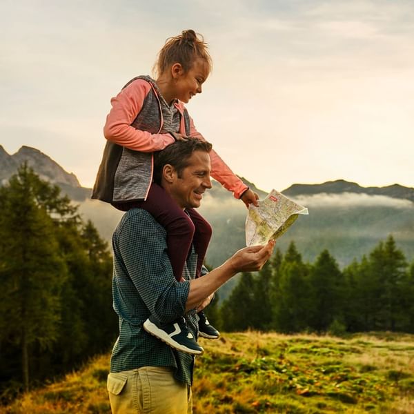 Vater mit Kind auf den Schultern, das eine Karte hält, vor Wald und Bergen.