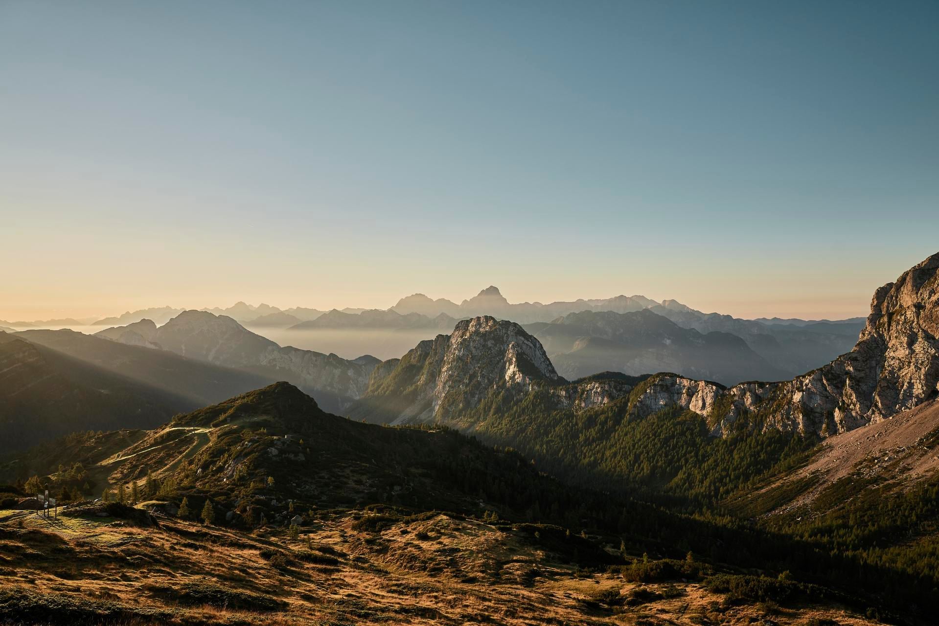 Bergige Landschaft mit Sonnenstrahlen und klarem Himmel im Hintergrund, umgeben von dichten Wäldern.