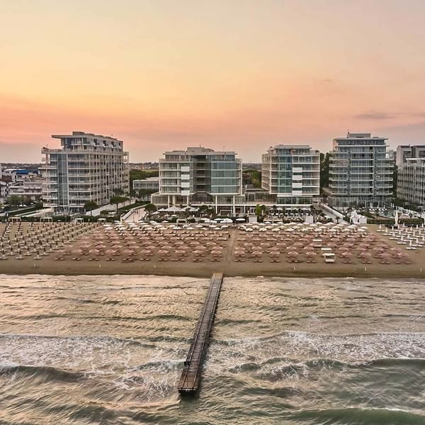 Strand mit vielen Schirmen und Liegen, umgeben von hohen Gebäuden und einer Holzbrücke im Wasser.