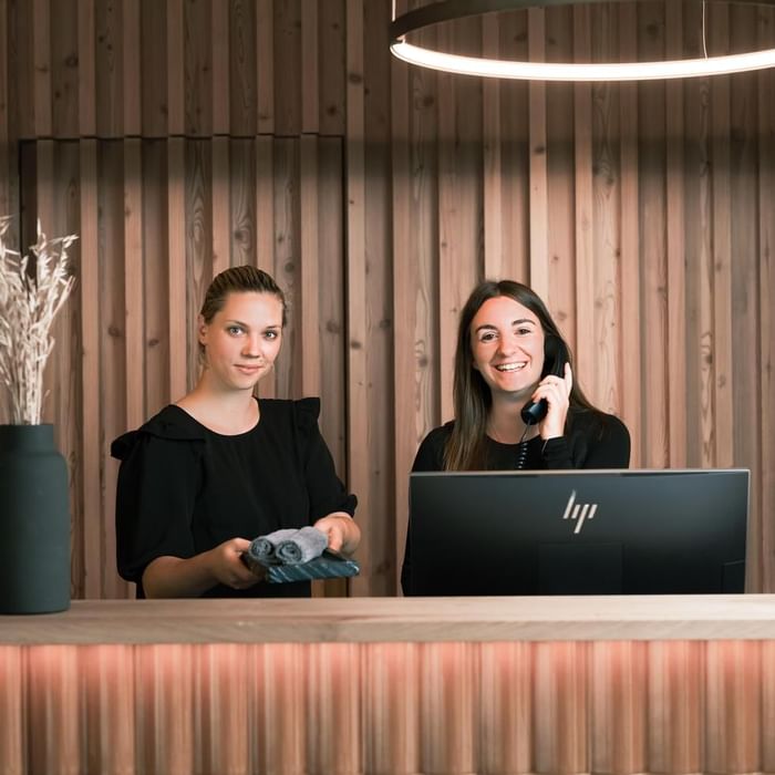 Zwei Frauen stehen hinter einer Rezeption, eine telefoniert, die andere hält Handtücher, vor ihnen ein Monitor.
