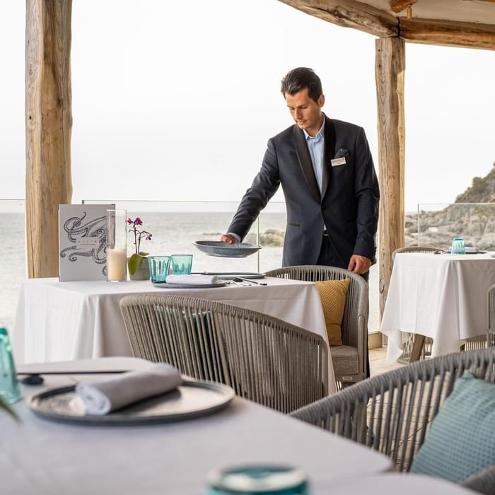 Mann in Anzug stellt Teller auf Tisch in einem Strandrestaurant mit Meerblick.