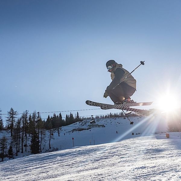 Get AI suggestions Skifahrer in der Luft über einer schneebedeckten Piste, umgeben von Bäumen und hellem Sonnenlicht.