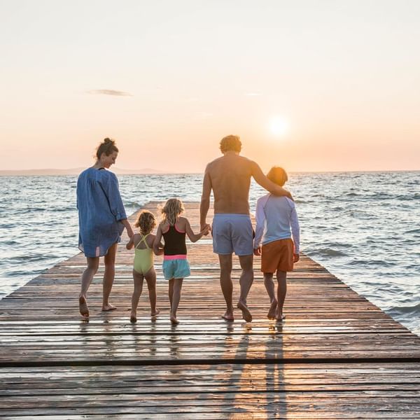 Familie geht am Abend auf einem Steg in Richtung Meer, während die Sonne untergeht.