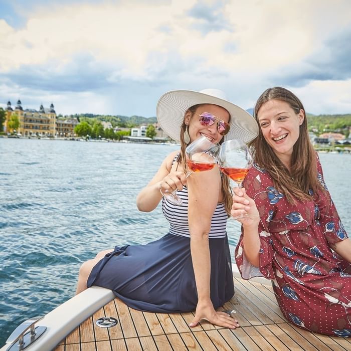 Zwei Frauen auf einer Bootsbank, die Wein trinken und lächeln, mit einer Stadt im Hintergrund.
