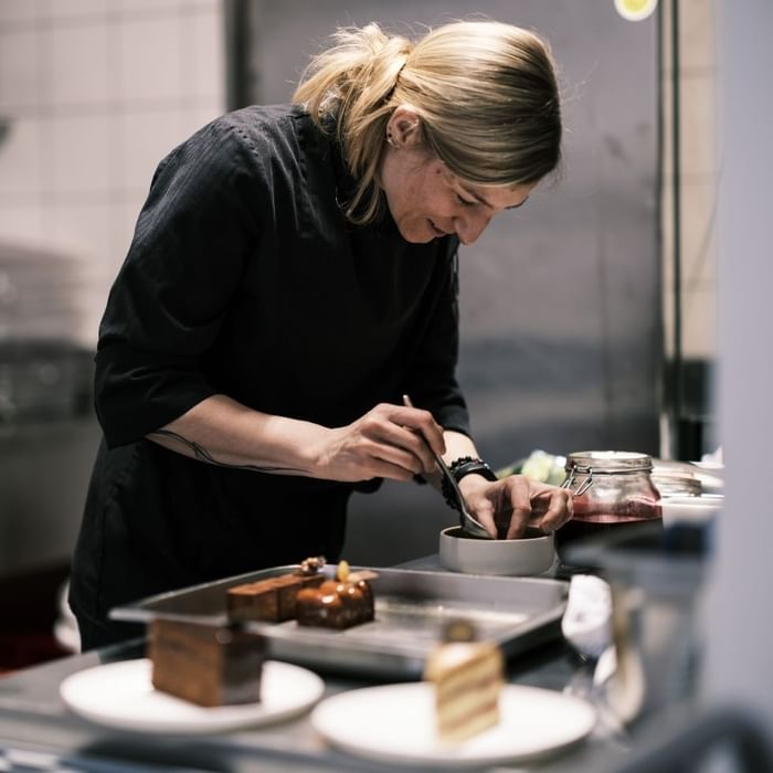 Blondhaarige Frau in schwarzer Kleidung bereitet Desserts in einer Küche vor.