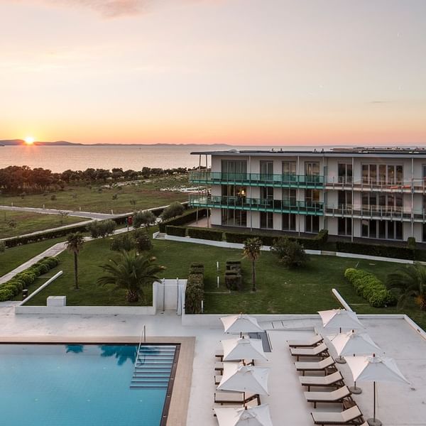 Sonnenuntergang über Hotelgebäude, Pool und Liegen, umgeben von Rasen und Bäumen.