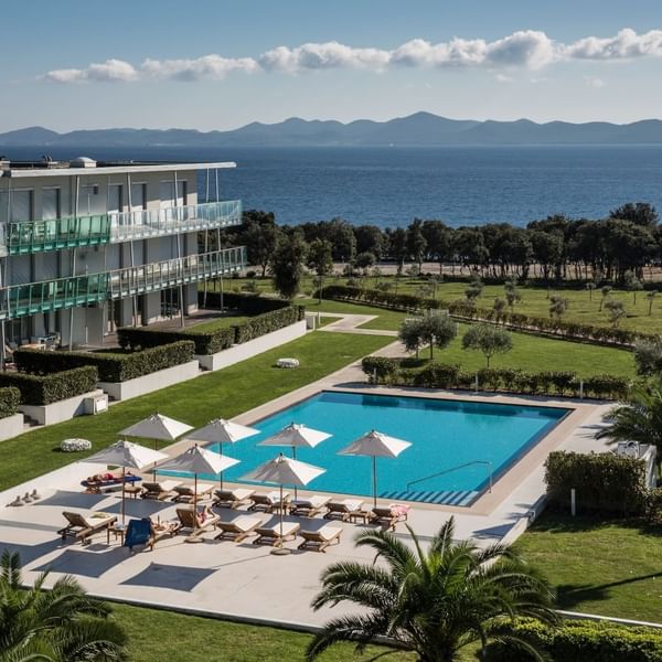 Hotelgebäude mit Meerblick, Pool und Sonnenschirmen, umgeben von Grün und Bäumen.