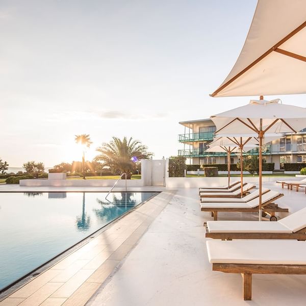 Pool mit Liegen und Sonnenschirmen neben modernem Gebäude unter hellem Himmel.