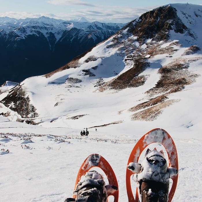 Zwei Schneeschuhe auf schneebedecktem Berg, mit Personen im Hintergrund und weitläufiger Berglandschaft.