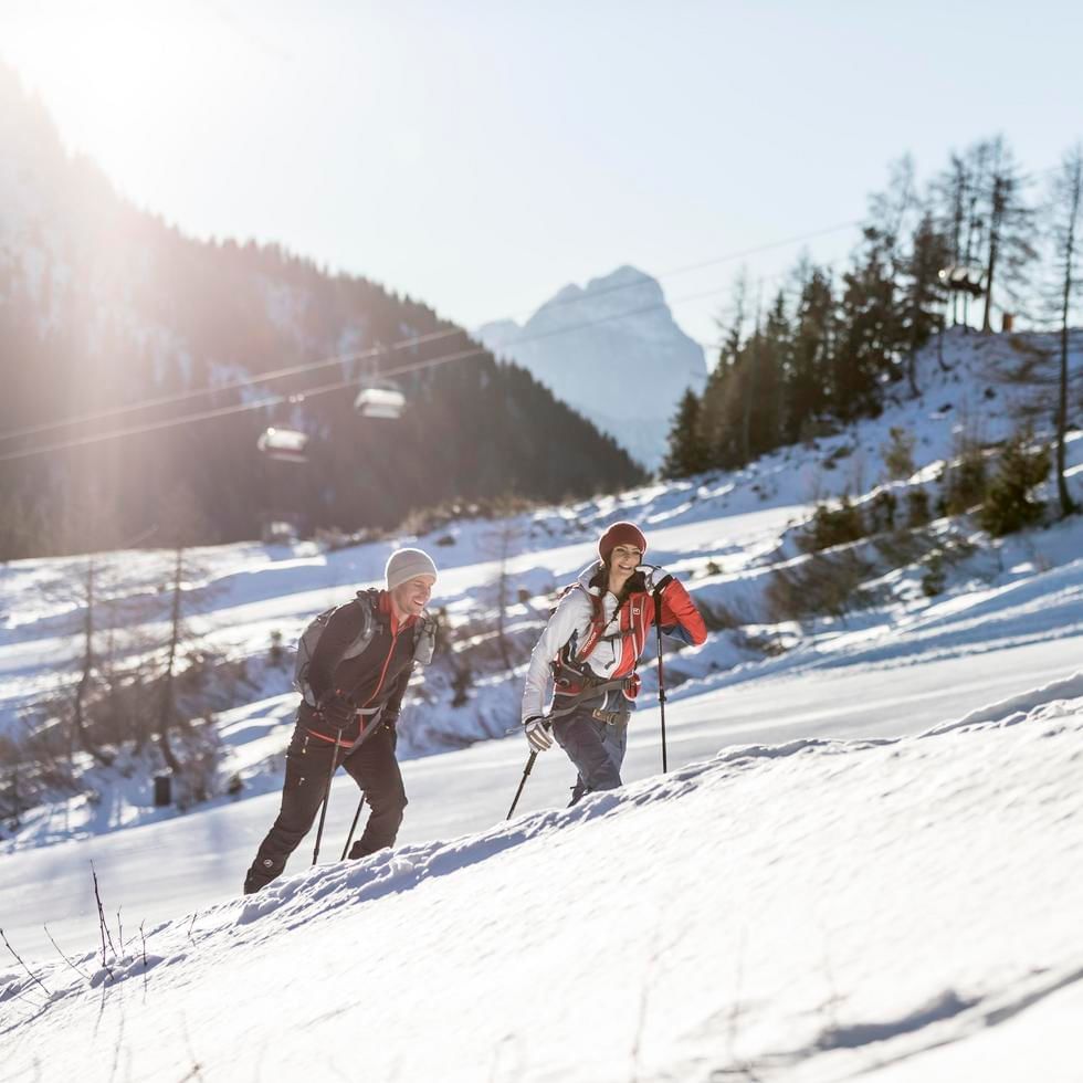 Zwei Wanderer mit Stöcken und Rucksäcken auf einem schneebedeckten Bergpfad, umgeben von winterlicher Natur.