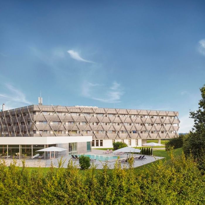 Modernes Hotelgebäude mit Poolbereich und Liegestühlen unter Sonnenschirmen, umgeben von Grün.