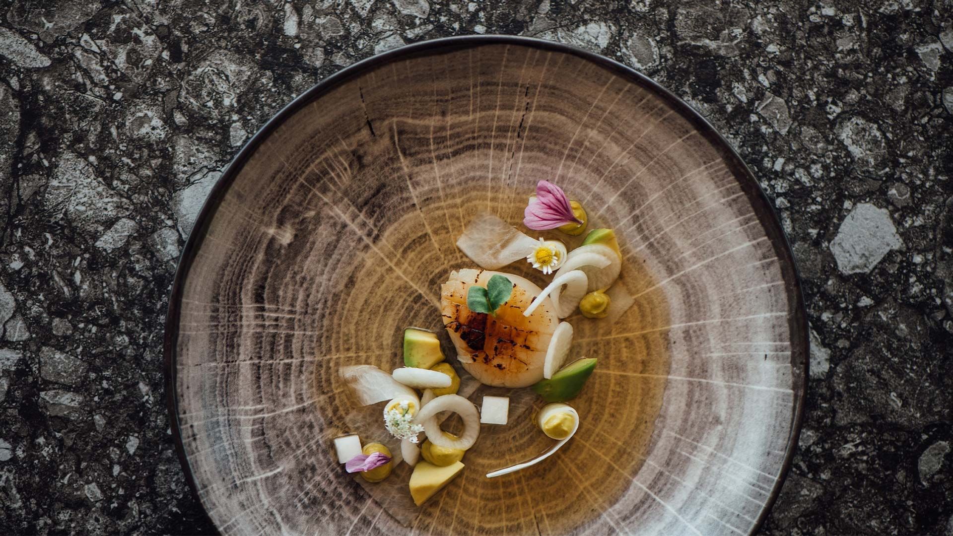 Piatto di pesce con verdure, avocado e fiori su un piatto circolare su una superficie di pietra.