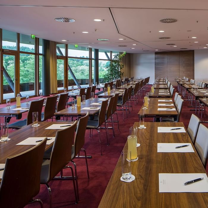 Konferenzraum mit vielen Tischen und Stühlen, Gläsern, Stiften und Unterlagen, umgeben von Fenstern und roter Teppich.