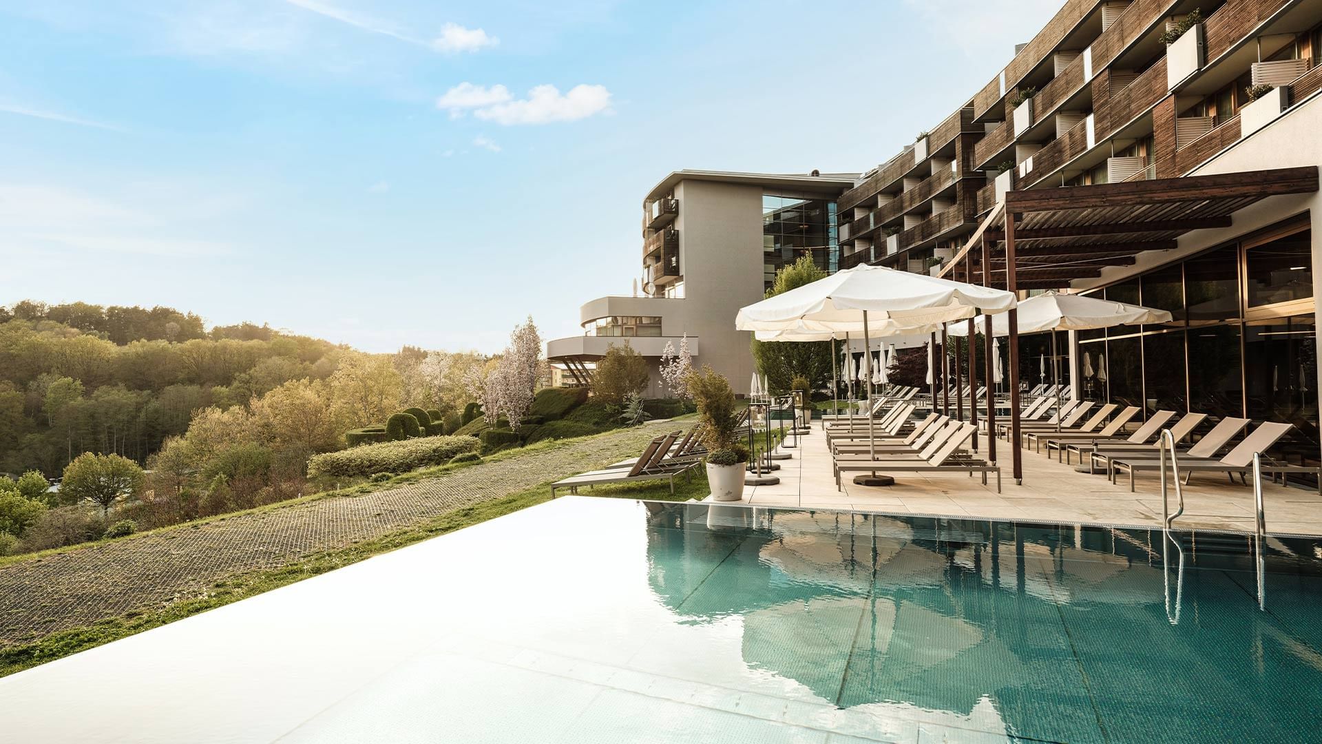 Piscina all'aperto con lettini e ombrelloni, affacciata su un'area verde e edifici moderni.