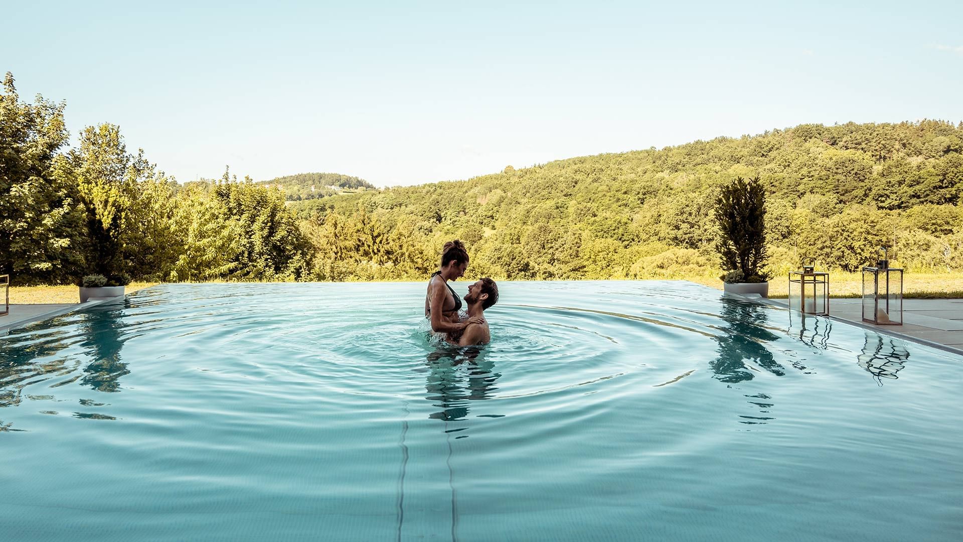 Coppia in piscina all'aperto con vista sulle colline boschive sotto un cielo blu.