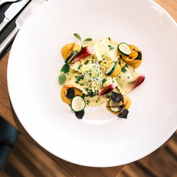 Piatto bianco con raviolo ripieno di zucca e zucchine, decorato con verdure e erbe aromatiche.