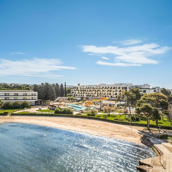 Luftaufnahme eines Resorts mit Strand, Pool, Gebäuden und viel Grün unter einem blauen Himmel.