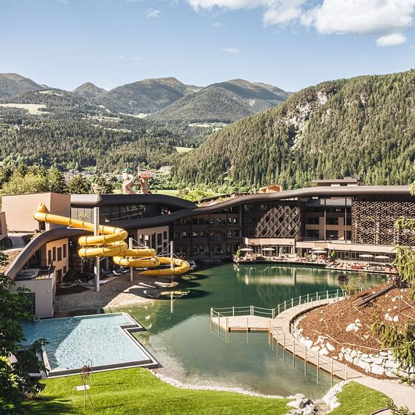Blick auf ein modernes Wellness-Resort mit gelber Wasserrutsche, Schwimmbecken und Bergkulisse.