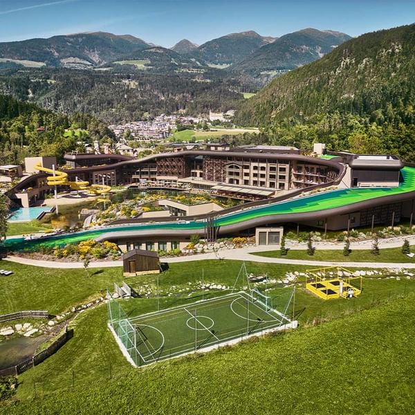 Luftaufnahme eines modernen Hotels mit Wasserrutschen, Pools und Sportfeldern in einer alpinen Landschaft.
