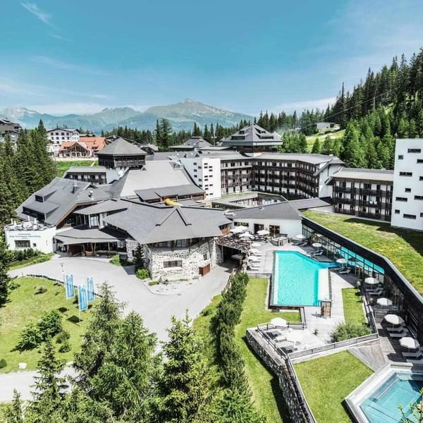 Luftaufnahme eines Hotelkomplexes mit Swimmingpool und umgeben von Wäldern und Bergen.