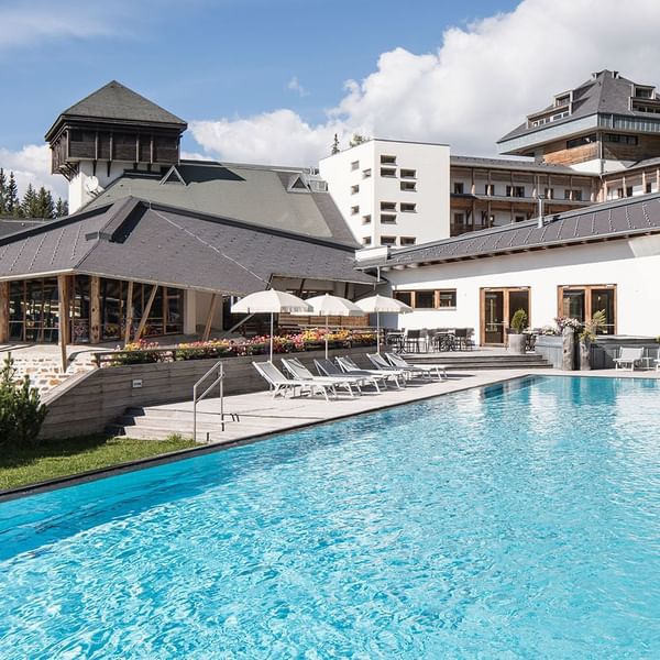 Großer Pool vor modernen Gebäuden mit Liegestühlen und Sonnenschirmen an einem sonnigen Tag.