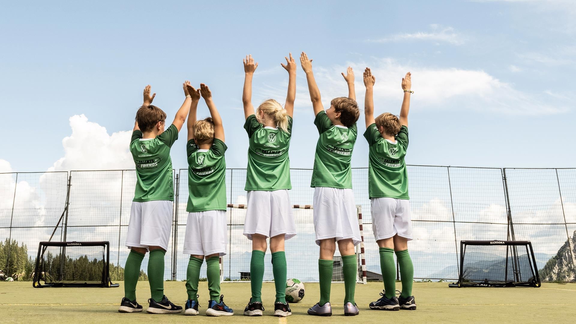 Sechs Kinder im Fußballtrikot mit erhobenen Armen vor dem Tor auf einem Sportplatz.