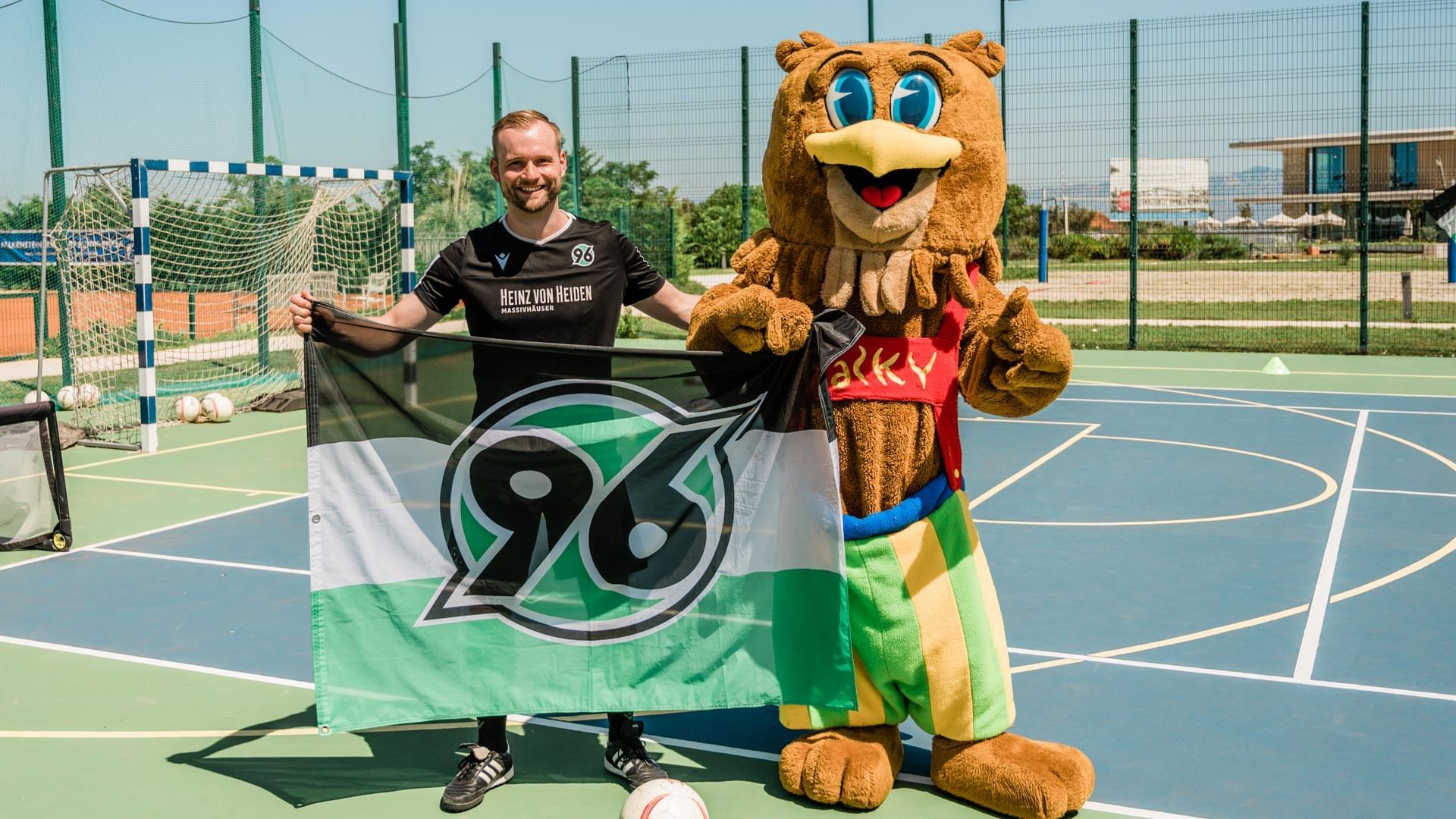 Mann mit Maskottchen und Fahne auf Sportplatz vor Fußballtor, umgeben von Bäumen und Gebäuden.
