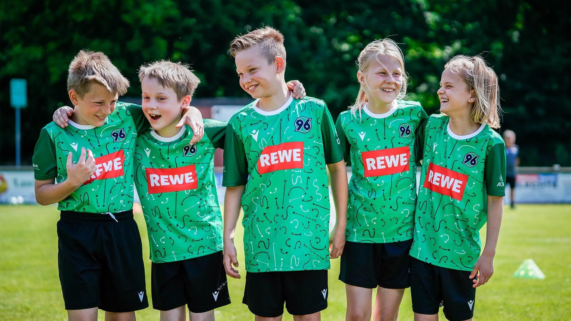 Fünf Kinder in REWE-Trikots stehen auf einem Rasen und lächeln für ein Foto.