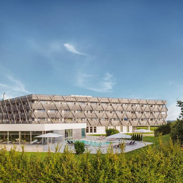 Modernes Hotelgebäude mit Außenpool und Liegestühlen unter weißen Schirmen, umgeben von Grün.