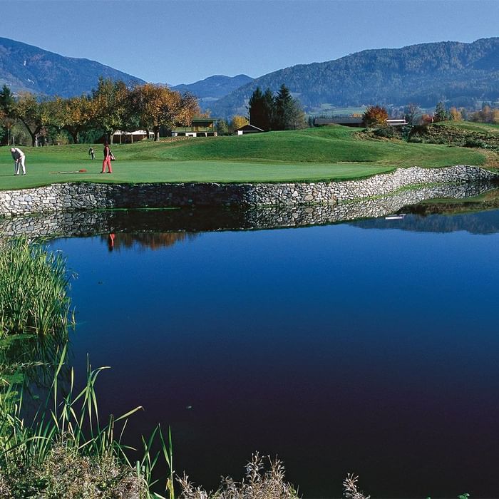 Golfplatz mit Teich und Spielern, umgeben von Bäumen und Bergen im Hintergrund.