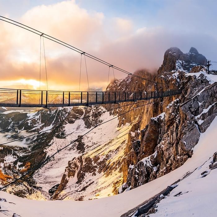 Hängebrücke über schneebedeckte Berge bei Sonnenuntergang mit Wolken und felsigen Gipfeln im Hintergrund.