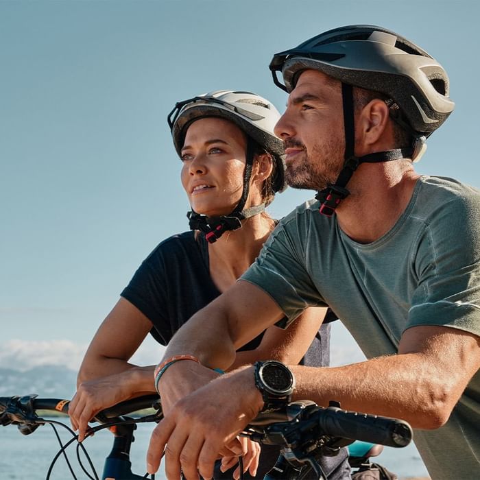 Zwei Radfahrer mit Helmen und Uhren sitzen auf Fahrrädern, nahe dem Meer.