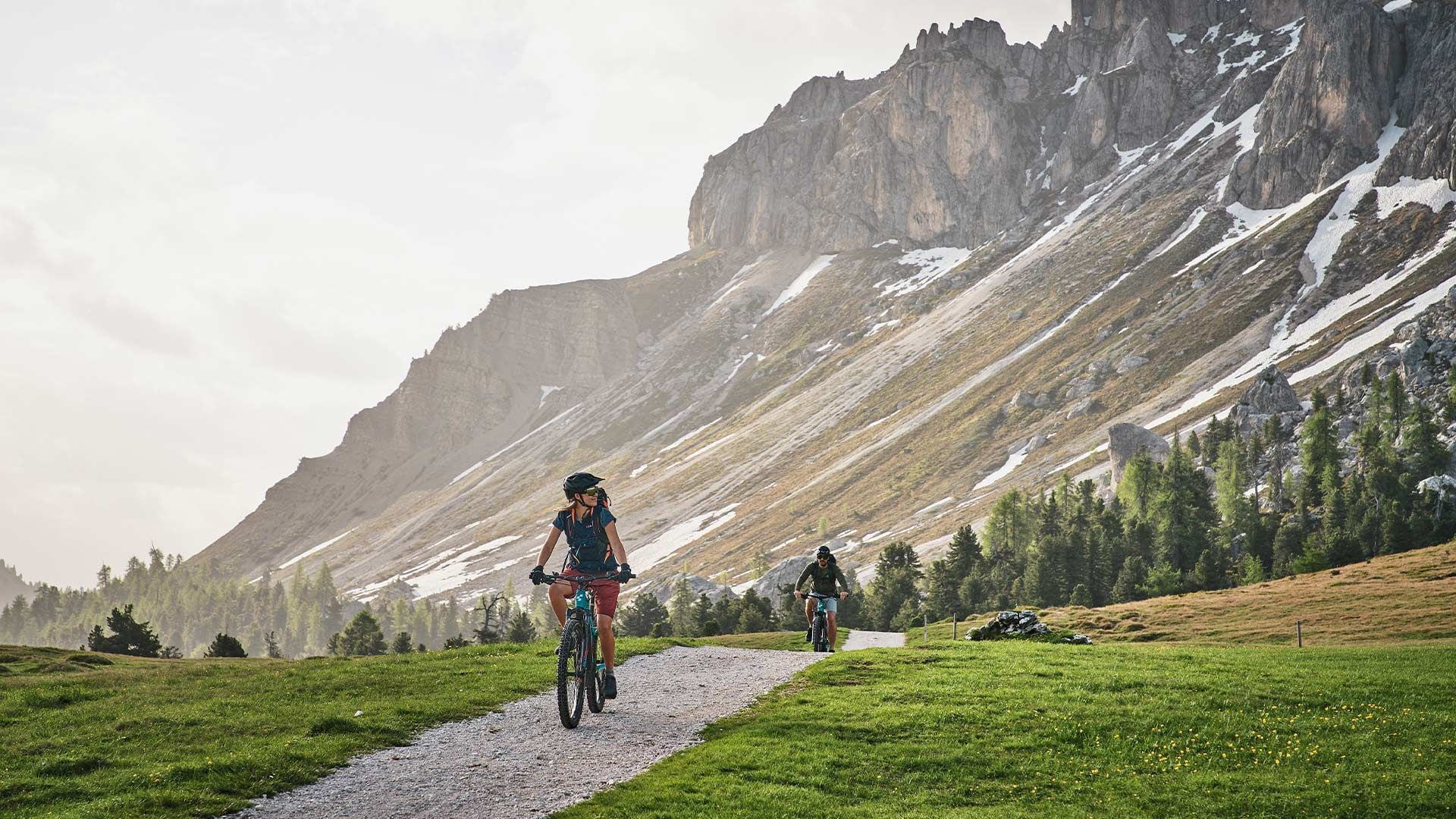Zwei Radfahrer fahren auf einem Pfad in einer alpinen Landschaft mit Schnee und Gras.
