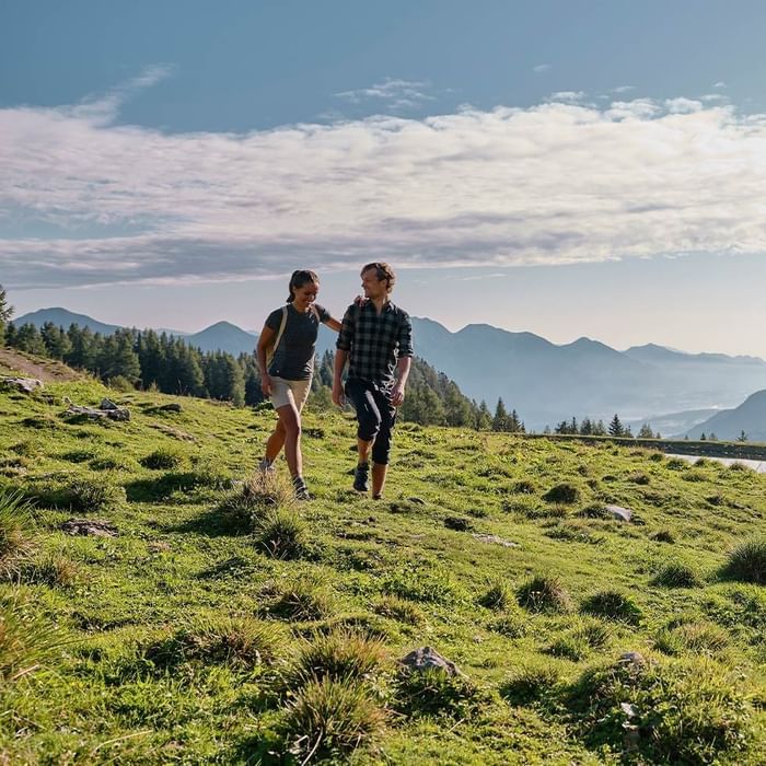 Zwei Personen wandern Hand in Hand auf einem grünen Bergpfad mit Bergpanorama im Hintergrund.