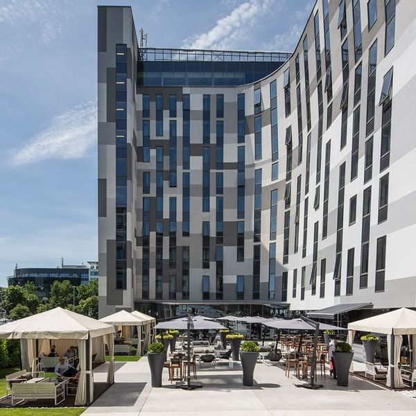 Modernes Gebäude mit vielen Fenstern und einer Terrasse mit Tischen, Stühlen und Schirmen davor.