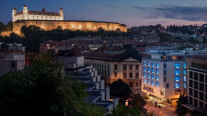 Abendliche Stadtansicht mit beleuchtetem Schloss und modernen Gebäuden, umgeben von Bäumen und Straßenbeleuchtung.