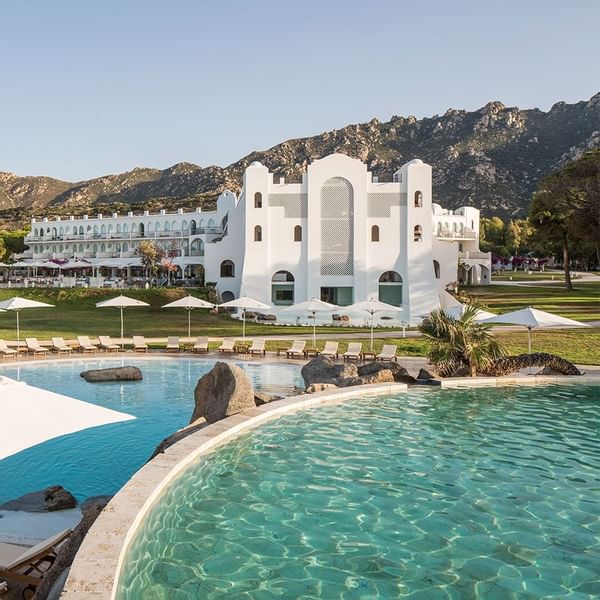 Großer Pool vor einem weißen Resorthotel mit vielen Liegen und Sonnenschirmen, umgeben von Bergen.