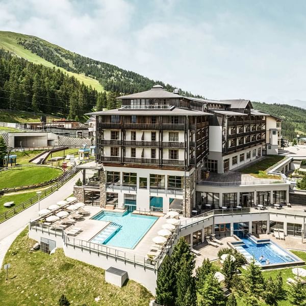 Luftaufnahme eines modernen Hotels mit zwei Pools, umgeben von bewaldeten Hügeln und Berglandschaft.