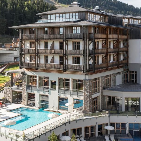 Modernes Hotelgebäude mit mehreren Etagen, Pool und Loungebereich, umgeben von einer bewaldeten Berglandschaft.