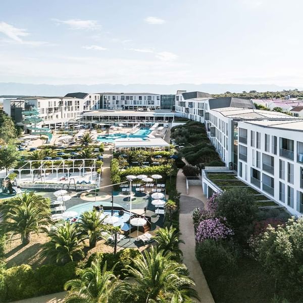 Luftaufnahme eines modernen Resorts mit Pools, Gebäuden, Gärten und Spazierwegen an einem sonnigen Tag.