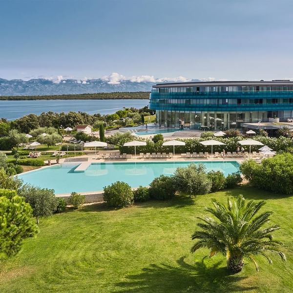 Luftaufnahme eines Resorts mit Pool, Liegen, Gebäuden und Meeresblick im Hintergrund.