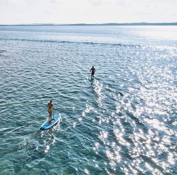 Zwei Personen paddeln auf Surfbrettern im kristallklaren Wasser einer malerischen Küstenbucht.