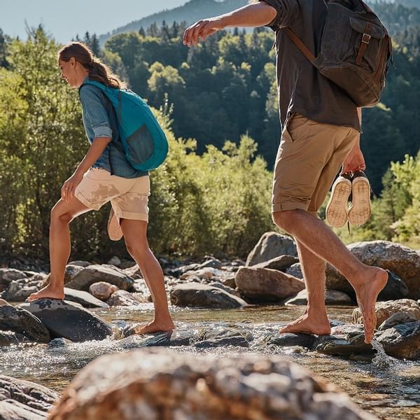 Mann und Frau wandern barfuß über Steine in einem Bach, umgeben von Wald und Bergen.