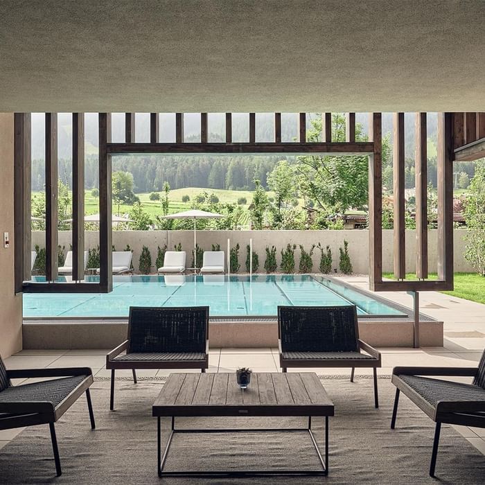 Innenbereich mit Lounge-Stühlen und Tisch vor einem Schwimmbad mit Blick auf grüne Landschaft und Berge.