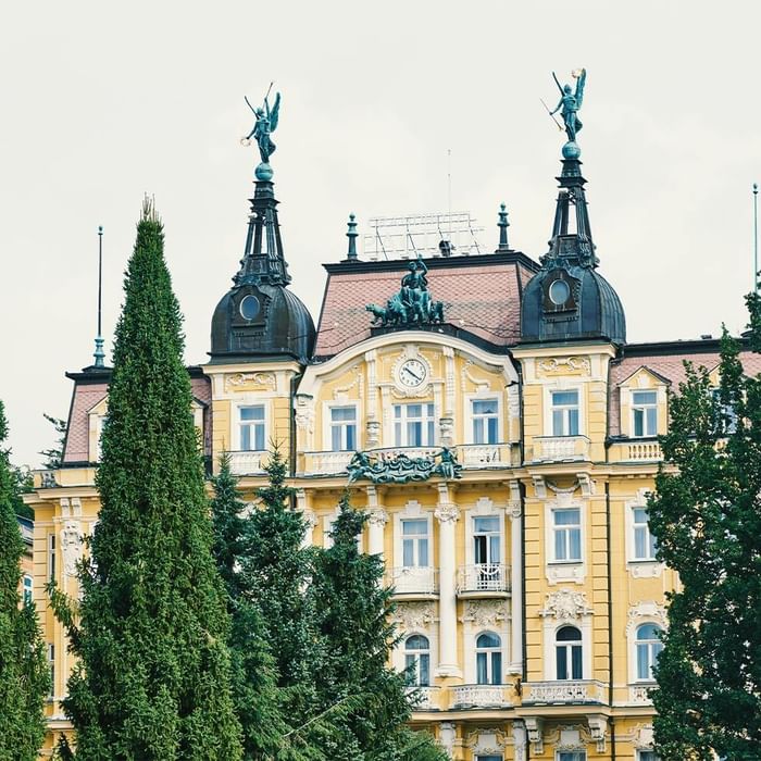 Gelbes Gebäude mit zwei Türmen und Statuen, umgeben von Bäumen und mit sichtbarer Uhr.