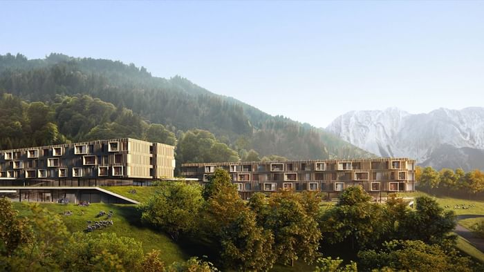 Modernes Gebäudekomplex in den Alpen mit vielen Fenstern, umgeben von Bäumen und grasenden Tieren.