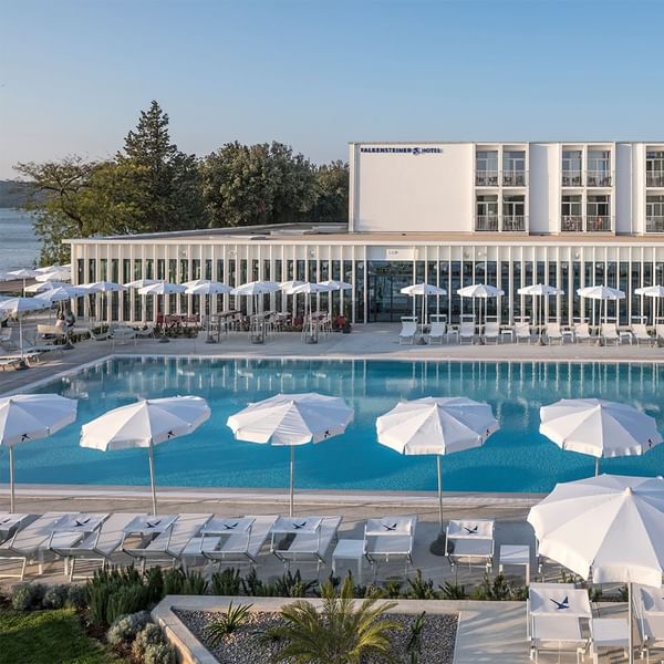 Luftaufnahme eines Hotelpools mit Liegestühlen, Sonnenschirmen und dem Hotelgebäude im Hintergrund.
