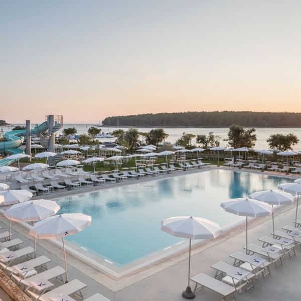 Poolbereich mit Liegen, Sonnenschirmen und Wasserrutsche vor der Küste bei Sonnenuntergang.